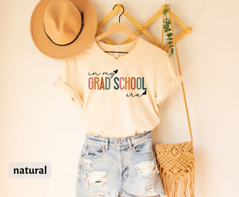 May include: A beige t-shirt with the text "in my grad school era" in rainbow colors and black butterflies. The shirt is hanging on a wooden hanger with a straw bag and a brown hat.