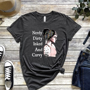 Op de afbeelding: Een donkergrijs T-shirt met een afbeelding van een vrouw met een boekenkast als hoofd. De tekst op het shirt luidt "Nerdy Dirty Inked And Curvy".