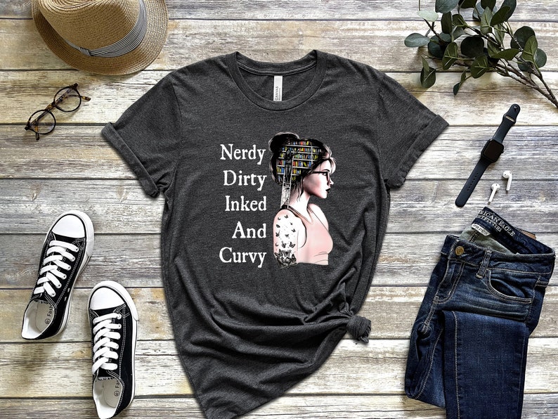 Op de afbeelding: Een donkergrijs T-shirt met een afbeelding van een vrouw met een boekenkast als hoofd. De tekst op het shirt luidt "Nerdy Dirty Inked And Curvy".