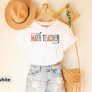 May include: White t-shirt with the text "in my MATH TEACHER era" in colourful letters. The shirt is hanging on a wooden rack with a straw bag and a potted plant.