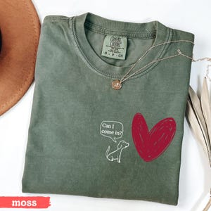 May include: Moss green t-shirt with a white cartoon dog and a red heart graphic. The text "Can I come in?" is in a speech bubble. A gold necklace is draped across the shirt.