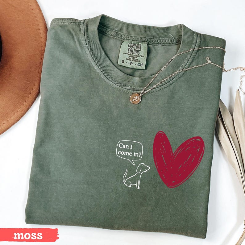 May include: Moss green t-shirt with a white cartoon dog and a red heart graphic. The text "Can I come in?" is in a speech bubble. A gold necklace is draped across the shirt.