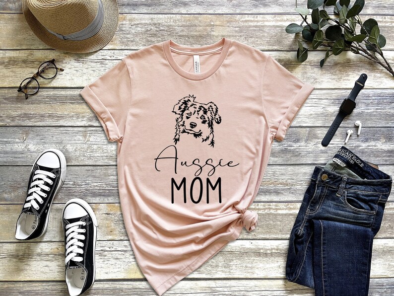 K&ouml;nnte beinhalten: Ein rosa T-Shirt mit einer schwarzen Umrandung eines Australian Shepherd Hundes und dem Text "Aussie MOM" auf der Vorderseite.