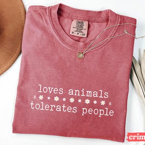 May include: A red t-shirt with the text "loves animals tolerates people" and a row of white flowers printed on the front. The shirt is made of a soft, comfortable material.