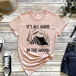May include: A pink t-shirt with a black graphic of a tent, mountains, and trees. The text "It's all good in the woods" is printed on the shirt.