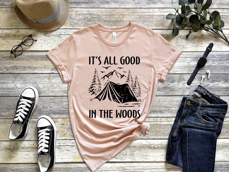 May include: A pink t-shirt with a black graphic of a tent, mountains, and trees. The text "It's all good in the woods" is printed on the shirt.