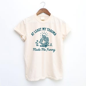 K&ouml;nnte beinhalten: Cremefarbenes T-Shirt mit dem t&uuml;rkisfarbenen Text "AT LEAST MY TRAUMA Made Me Funny". Die Grafik zeigt ein Skelett, das im Schneidersitz sitzt und eine Tasse h&auml;lt, mit Blumen. Das T-Shirt h&auml;ngt an einem Holzb&uuml;gel.