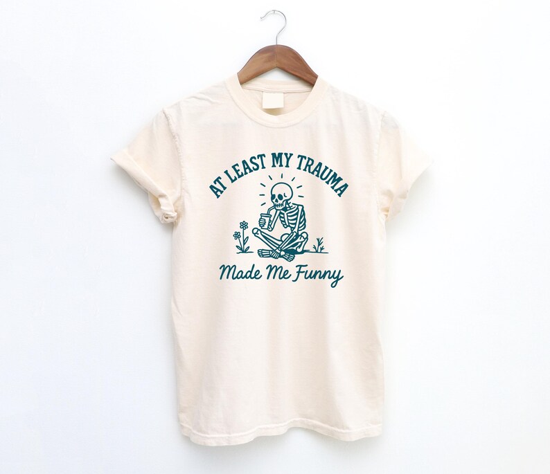 K&ouml;nnte beinhalten: Cremefarbenes T-Shirt mit dem t&uuml;rkisfarbenen Text "AT LEAST MY TRAUMA Made Me Funny". Die Grafik zeigt ein Skelett, das im Schneidersitz sitzt und eine Tasse h&auml;lt, mit Blumen. Das T-Shirt h&auml;ngt an einem Holzb&uuml;gel.