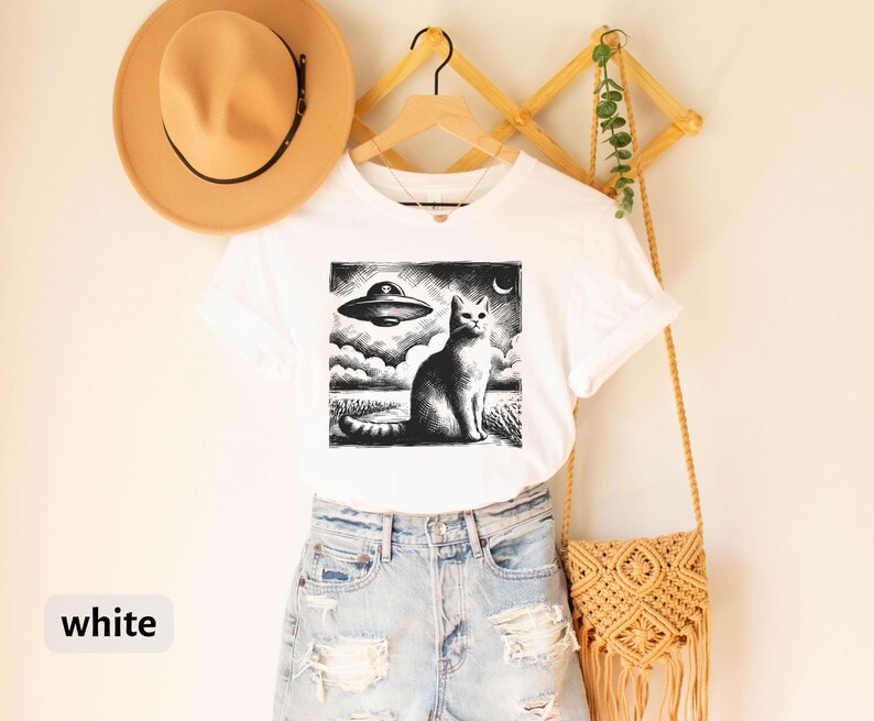 May include: White t-shirt with a black and white graphic print of a cat looking up at a UFO. The cat is sitting on a field with a crescent moon in the sky. The shirt is on a hanger with a straw hat and a woven bag.