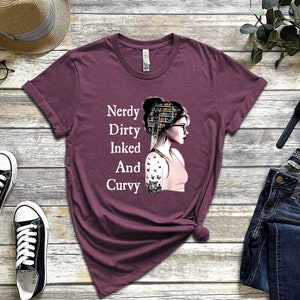 Op de afbeelding: Een maroon t-shirt met de tekst "Nerdy Dirty Inked And Curvy" en een afbeelding van een vrouw met boeken in haar haar en een tatoeage op haar arm.