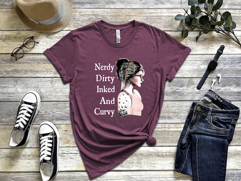 Op de afbeelding: Een maroon t-shirt met de tekst "Nerdy Dirty Inked And Curvy" en een afbeelding van een vrouw met boeken in haar haar en een tatoeage op haar arm.