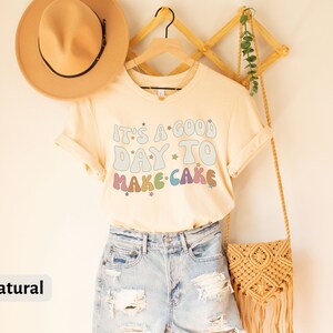 Peut inclure: Un t-shirt beige clair avec le texte "It's a good day to make cake" en couleurs arc-en-ciel avec des &eacute;toiles. Le t-shirt est suspendu &agrave; un cintre en bois avec un sac en paille et un chapeau.