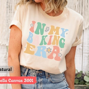 May include: A beige t-shirt with the text "In My Baking Era" in colorful retro-style lettering. The shirt has a butterfly and flower graphic.