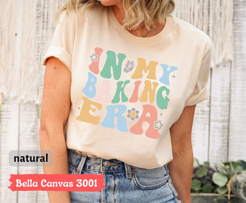 May include: A beige t-shirt with the text "In My Baking Era" in colorful retro-style lettering. The shirt has a butterfly and flower graphic.