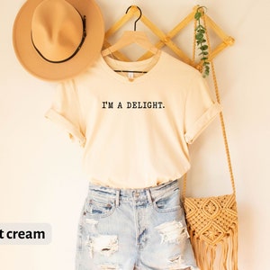 May include: A soft cream colored t-shirt with the text "I'M A DELIGHT." printed on the front. The t-shirt is paired with light blue denim shorts and a straw bag.
