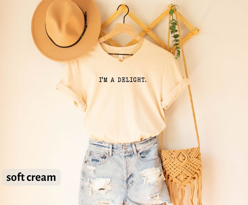 May include: A soft cream colored t-shirt with the text "I'M A DELIGHT." printed on the front. The t-shirt is paired with light blue denim shorts and a straw bag.