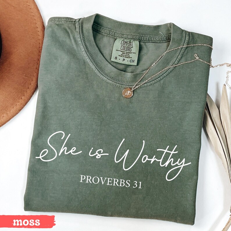 May include: A moss green Comfort Colors t-shirt with the words "She is Worthy" and "Proverbs 31" printed in white. A gold necklace with a pendant rests on the shirt. The shirt is folded and placed on a white surface.