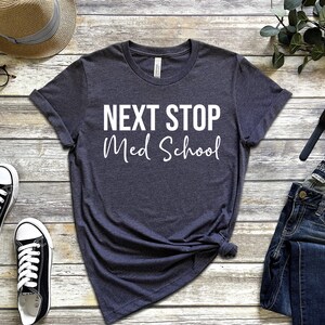 May include: A dark gray t-shirt with the text "NEXT STOP Med School" in white lettering. The shirt is laid out on a wooden surface with a hat, glasses, jeans, a watch, and earbuds.