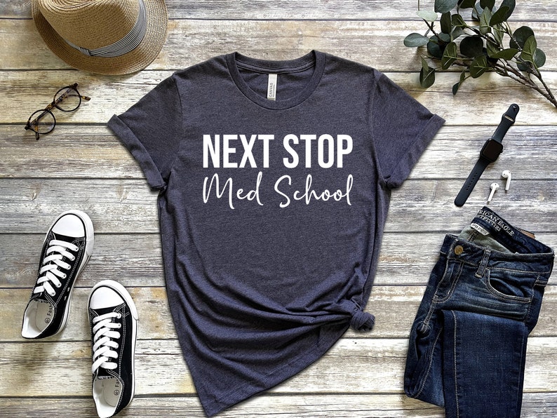 May include: A dark gray t-shirt with the text "NEXT STOP Med School" in white lettering. The shirt is laid out on a wooden surface with a hat, glasses, jeans, a watch, and earbuds.