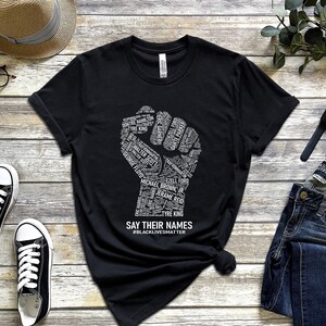 Puede incluir: Camiseta negra con un gr&aacute;fico de pu&ntilde;o blanco hecho de nombres. El gr&aacute;fico del pu&ntilde;o est&aacute; rodeado del texto "SAY THEIR NAMES #BLACKLIVESMATTER".