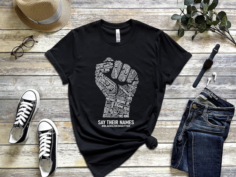 Puede incluir: Camiseta negra con un gr&aacute;fico de pu&ntilde;o blanco hecho de nombres. El gr&aacute;fico del pu&ntilde;o est&aacute; rodeado del texto "SAY THEIR NAMES #BLACKLIVESMATTER".
