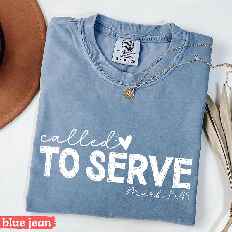 Pu&ograve; includere: T-shirt blu jeans con le parole "called to serve" in scrittura bianca e lettere maiuscole. Una collana d'oro con un ciondolo &egrave; appoggiata sulla maglietta. Il testo "Mark 10:45" &egrave; sotto. La maglietta &egrave; piegata.