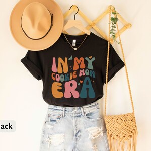 May include: Black t-shirt with the text "In My Cookie Mom Era" in colorful retro font. The shirt is hanging on a wooden rack with a straw bag and a hat.
