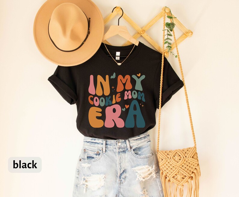 May include: Black t-shirt with the text "In My Cookie Mom Era" in colorful retro font. The shirt is hanging on a wooden rack with a straw bag and a hat.