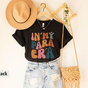 May include: Black t-shirt with the text "In My Para Era" in colorful, retro-style lettering. The shirt is hanging on a wooden rack with a straw hat and a woven bag.