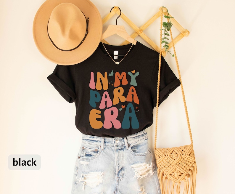 May include: Black t-shirt with the text "In My Para Era" in colorful, retro-style lettering. The shirt is hanging on a wooden rack with a straw hat and a woven bag.