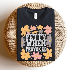 May include: Black t-shirt with the text "PETTY WHEN PROVOKED" in white, surrounded by colorful flowers in shades of yellow, pink, and purple. The shirt is displayed on a woven, circular surface.
