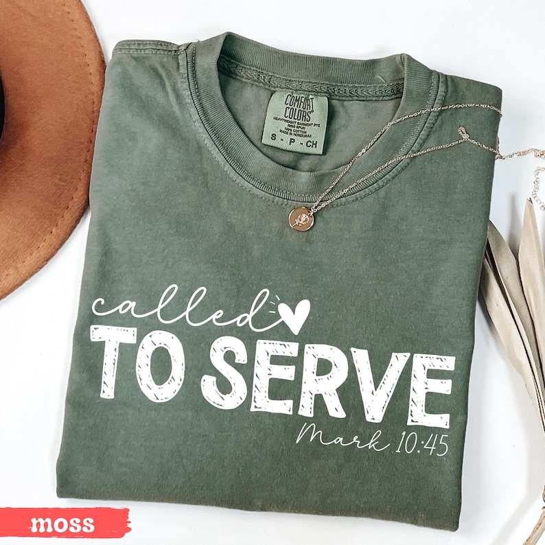 Pu&ograve; includere: Una maglietta verde muschio con le parole "called TO SERVE" in bianco, insieme a "Mark 10:45". Una collana d'oro con un ciondolo poggia sulla maglietta. La maglietta &egrave; piegata e ha un'etichetta che dice "Comfort Colors".