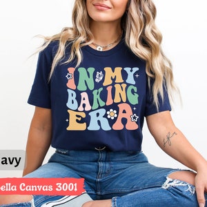 May include: A navy blue t-shirt with the text "In My Baking Era" in colorful retro lettering. The shirt features a butterfly and flower graphic.