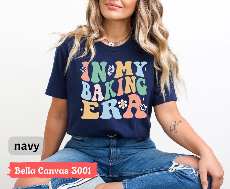 May include: A navy blue t-shirt with the text "In My Baking Era" in colorful retro lettering. The shirt features a butterfly and flower graphic.