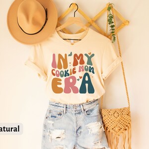 May include: A beige t-shirt with the text "In My Cookie Mom Era" in colorful, retro-style lettering. The shirt is on a hanger and has a natural color.