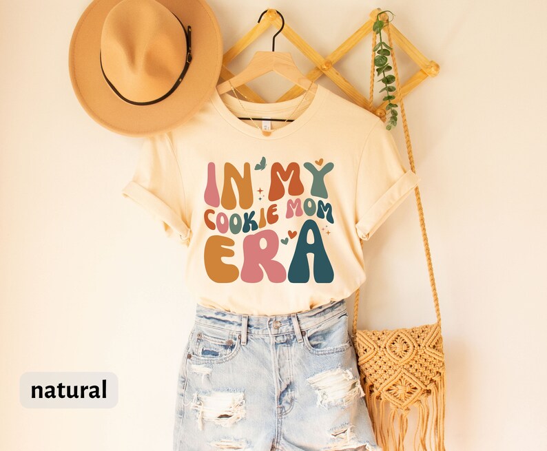 May include: A beige t-shirt with the text "In My Cookie Mom Era" in colorful, retro-style lettering. The shirt is on a hanger and has a natural color.