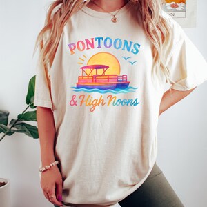 May include: Cream-colored t-shirt with a colorful graphic of a pontoon boat, sun, and birds. The text "Pontoons & High Noons" is printed above and below the boat in a rainbow gradient. The shirt is worn by a person.
