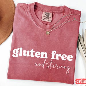 Puede incluir: Camiseta de color carmes&iacute; con las palabras "gluten free and starving" en blanco. Un collar plateado con un colgante descansa sobre la camiseta. La camiseta tiene cuello redondo y una etiqueta en la parte superior.