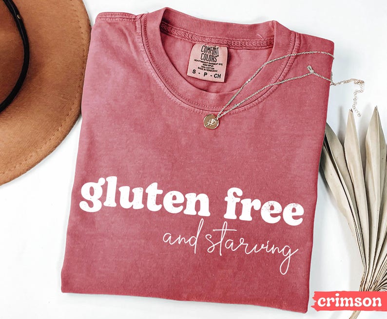 Puede incluir: Camiseta de color carmes&iacute; con las palabras "gluten free and starving" en blanco. Un collar plateado con un colgante descansa sobre la camiseta. La camiseta tiene cuello redondo y una etiqueta en la parte superior.