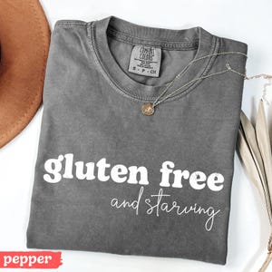 Puede incluir: Una camiseta gris con las palabras "gluten free and starving" en escritura blanca. La camiseta tiene cuello redondo y mangas cortas. Un collar dorado con un peque&ntilde;o colgante descansa sobre la camiseta. Un sombrero marr&oacute;n es visible en la esquina superior izquierda.