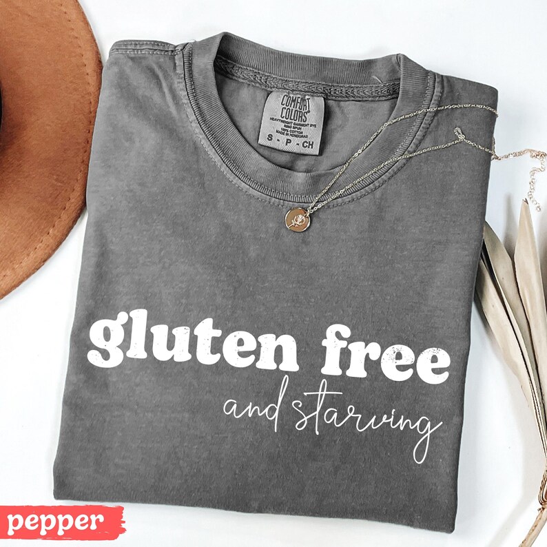 Puede incluir: Una camiseta gris con las palabras "gluten free and starving" en escritura blanca. La camiseta tiene cuello redondo y mangas cortas. Un collar dorado con un peque&ntilde;o colgante descansa sobre la camiseta. Un sombrero marr&oacute;n es visible en la esquina superior izquierda.