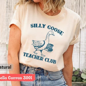 May include: A cream-colored t-shirt with the text "Silly Goose Teacher Club" in blue lettering. A cartoon goose carrying books is pictured below the text.