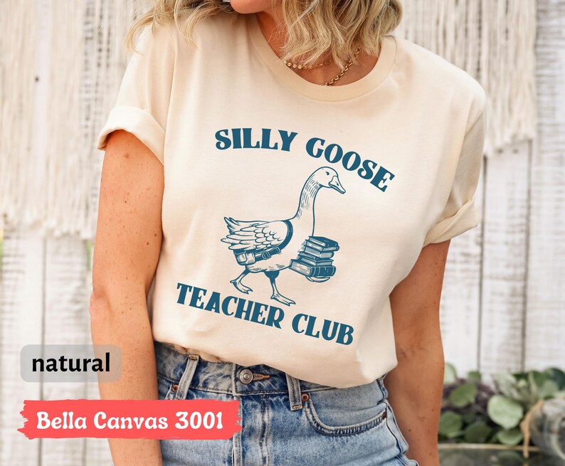 May include: A cream-colored t-shirt with the text "Silly Goose Teacher Club" in blue lettering. A cartoon goose carrying books is pictured below the text.