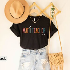 May include: Black t-shirt with the text "in my MATH TEACHER era" in rainbow colours. The shirt is on a hanger with a straw hat and a woven bag.