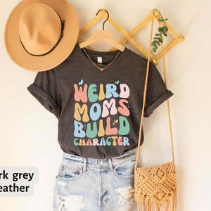 May include: Dark grey heather t-shirt with the text "Weird Moms Build Character" in colourful, retro-style lettering.