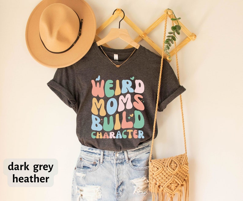 May include: Dark grey heather t-shirt with the text "Weird Moms Build Character" in colourful, retro-style lettering.