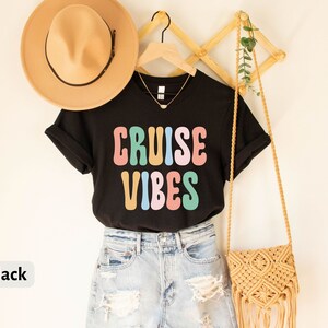 Puede incluir: Camiseta negra con el texto "CRUISE VIBES" en letras de colores de estilo retro. La camiseta se lleva con pantalones cortos de mezclilla y un bolso de paja.
