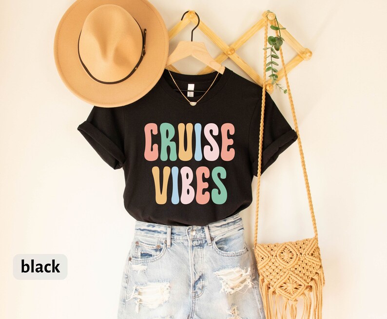 Puede incluir: Camiseta negra con el texto "CRUISE VIBES" en letras de colores de estilo retro. La camiseta se lleva con pantalones cortos de mezclilla y un bolso de paja.