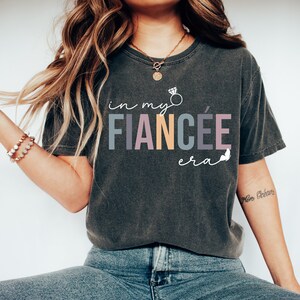 May include: A dark gray t-shirt with the text "in my fianc&eacute;e era" in colorful letters. A diamond ring is drawn above the word "fianc&eacute;e".