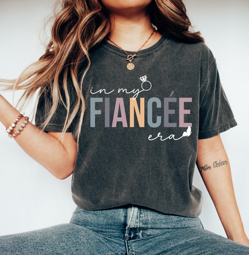 May include: A dark gray t-shirt with the text "in my fianc&eacute;e era" in colorful letters. A diamond ring is drawn above the word "fianc&eacute;e".